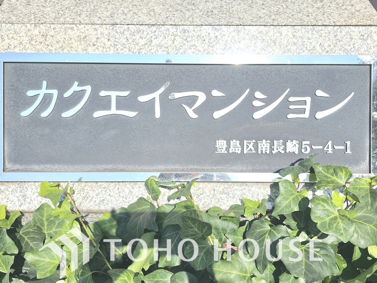 エントランス 「カクエイマンション」の文字が刻まれた、重厚感のある銘板です。洗練されたデザインが、住まいの顔として上品な印象を与えます。緑の植栽が彩りを添え、落ち着いた雰囲気を醸し出しています。